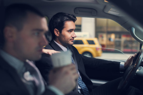 Young businessman driving a car in New York city to a business meeting with his passanger partnes who is dringing coffee. Horizontal shot inside a vehicle with visible yellow cab.